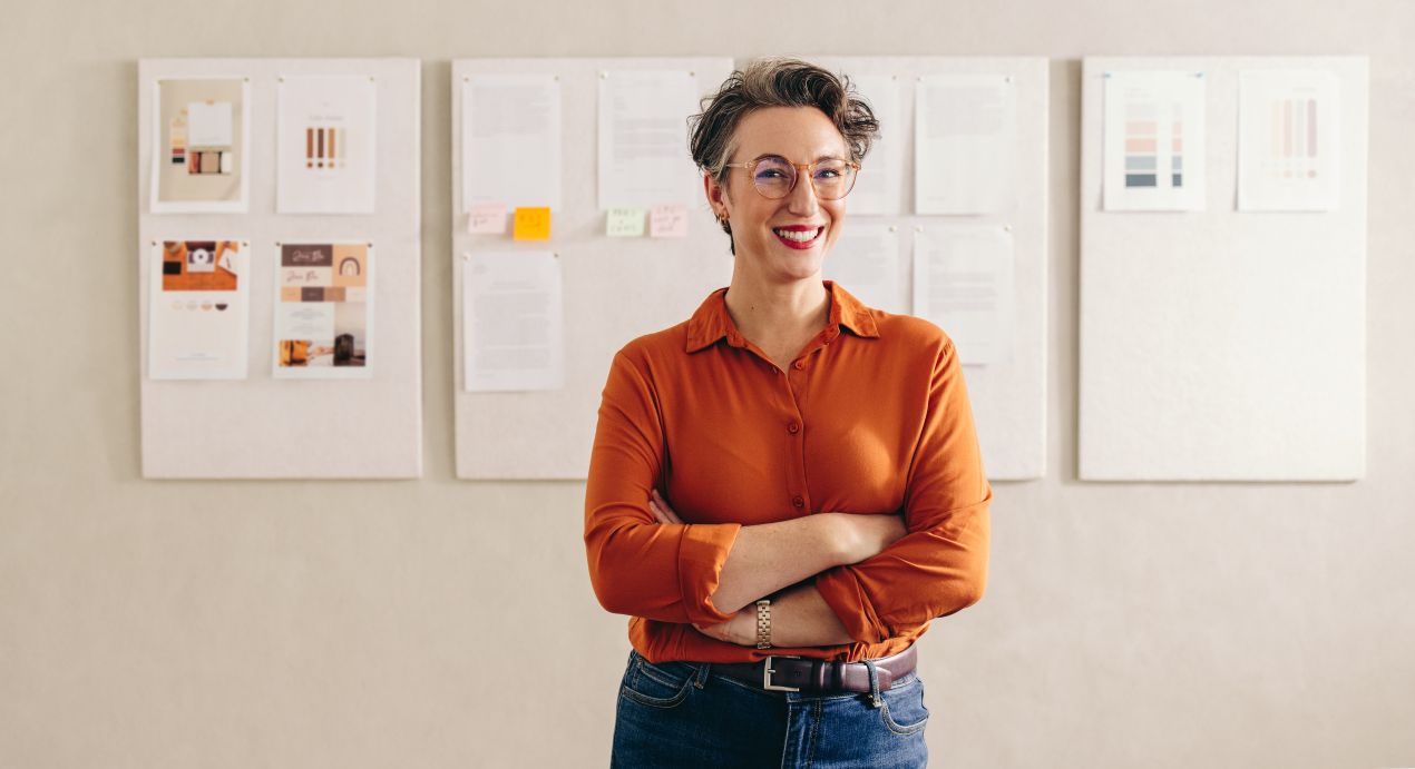 Interior-Designerin in orangefarbener Bluse steht mit verschränkten Armen vor einer Wand mit Moodboards, Farbmusterkarten und Notizen zur Wohnraumgestaltung.