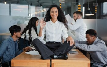 Eine lächelnde Frau sitzt im Schneidersitz auf einem Tisch und zeigt ein „Okay“-Zeichen, während ihr Team im Hintergrund arbeitet – Symbol für positive Teamkultur und mentale Gesundheit im Arbeitsumfeld.