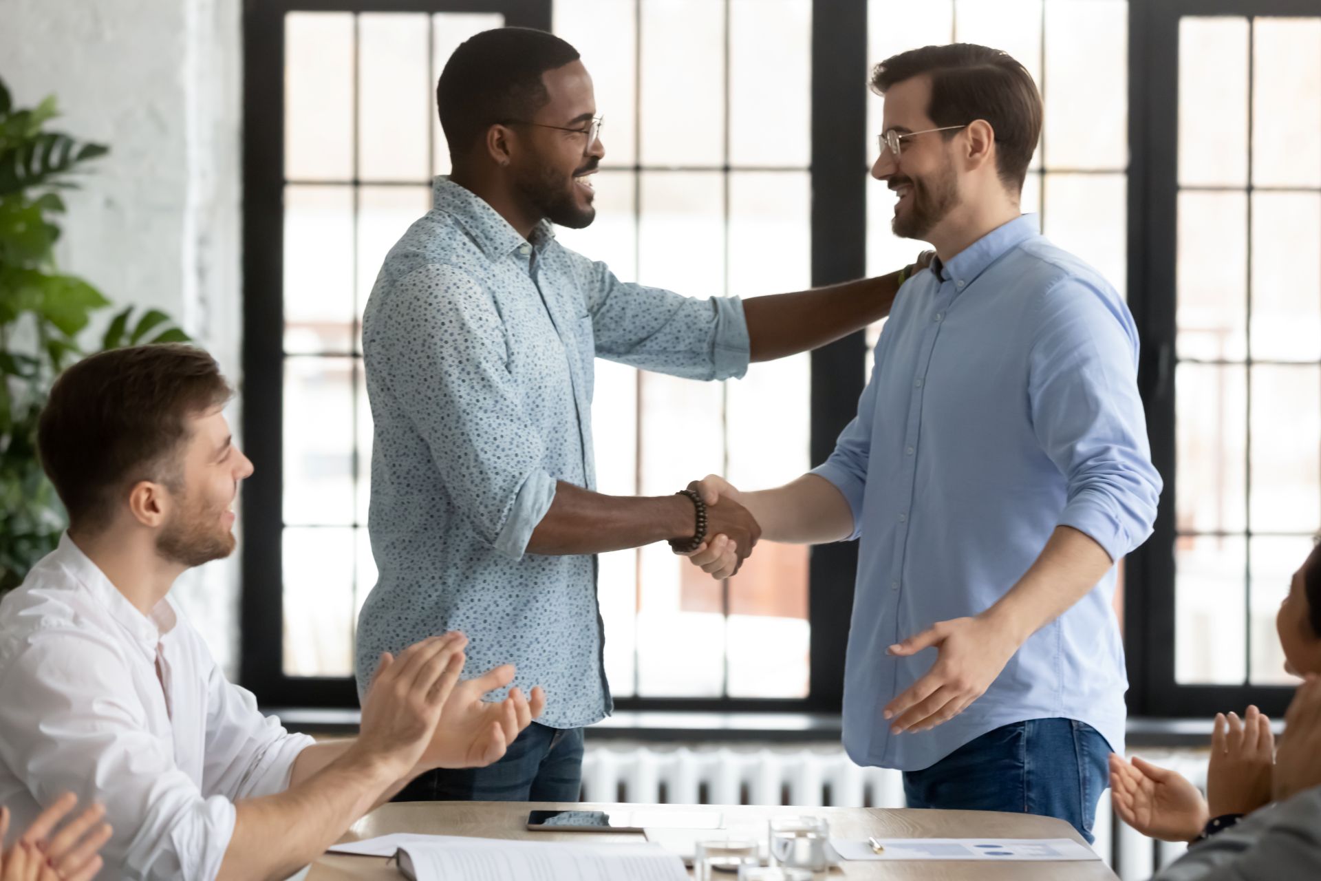 Zwei Männer geben sich die Hand und lächeln, während Kolleg:innen applaudieren – Symbol für Vertrauen, erfolgreiche Kommunikation und konstruktive Konfliktlösung im Unternehmen.  